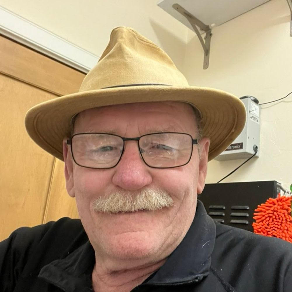 Older man with glasses and a mustache wearing a tan hat, smiling in a workshop setting with tools in the background.